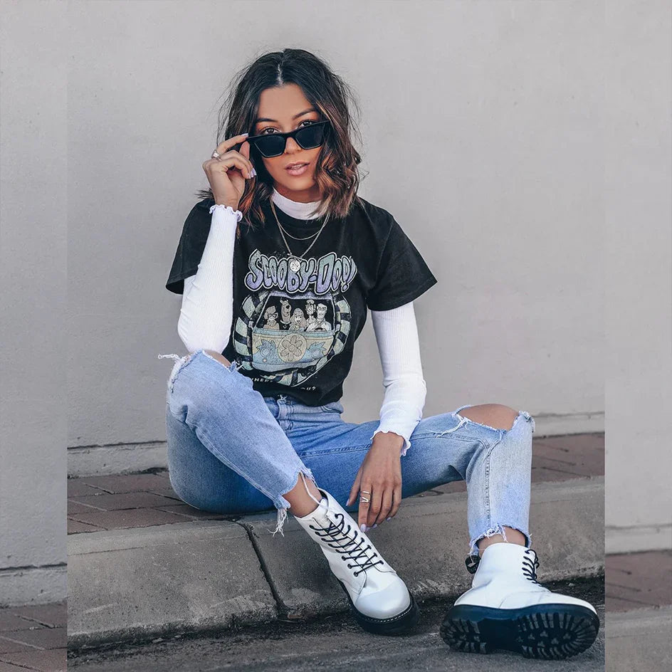 Woman in ripped jeans, graphic tee, white boots, and sunglasses sitting on a sidewalk