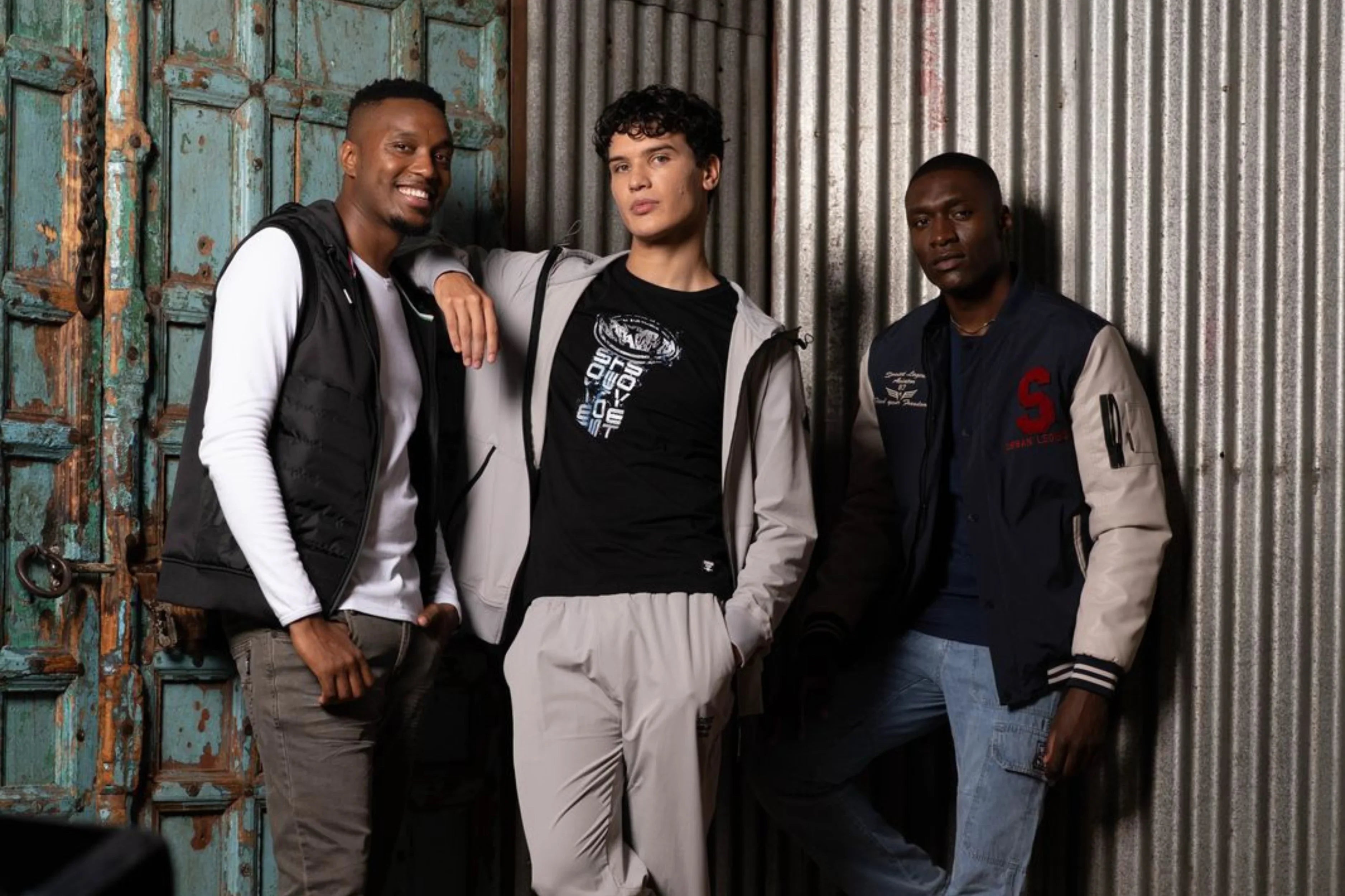 Three men in trendy urban streetwear pose against industrial metal and rustic wood doors.